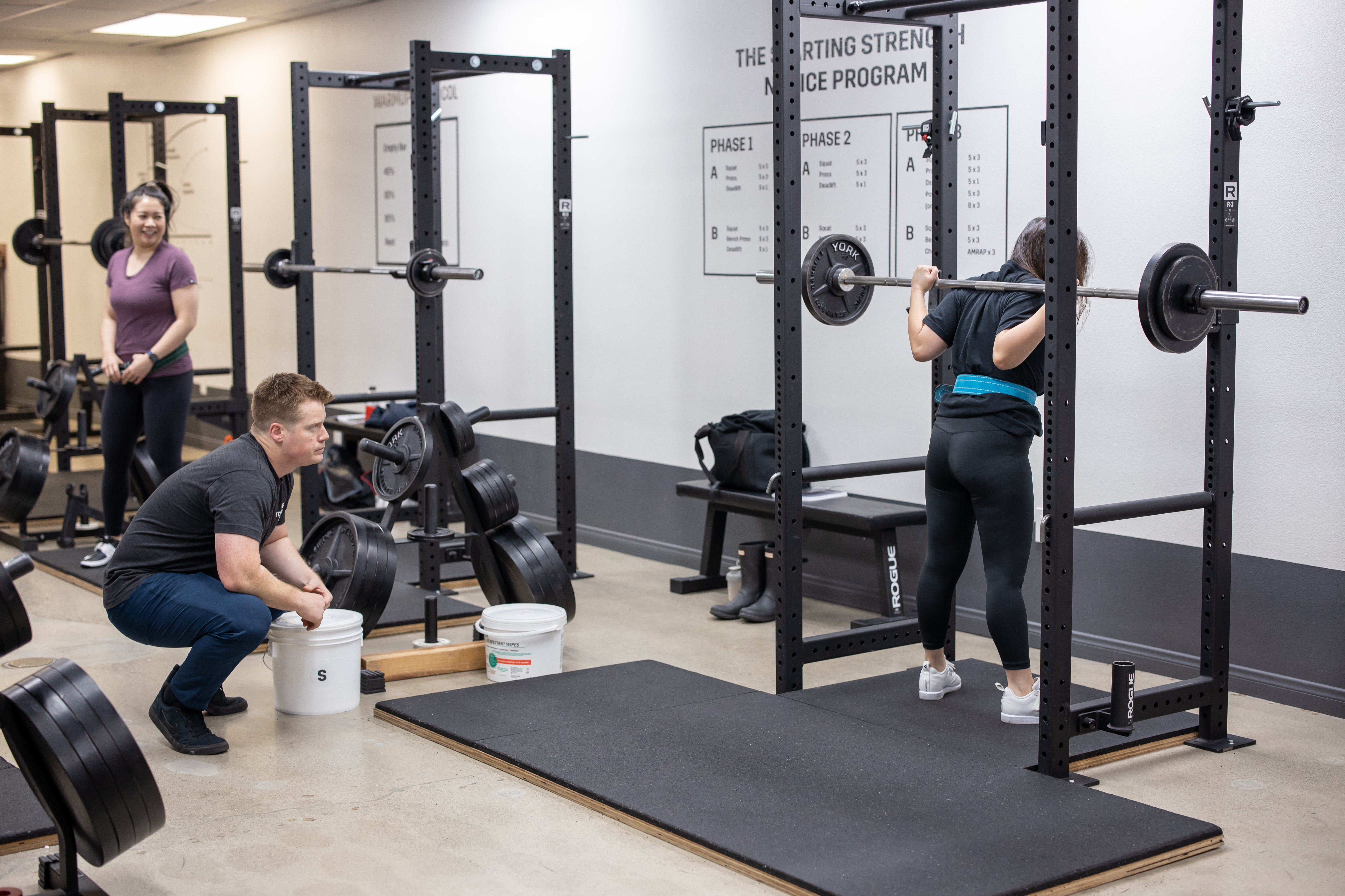 A woman squats in a squat rack while a coach looks on.
