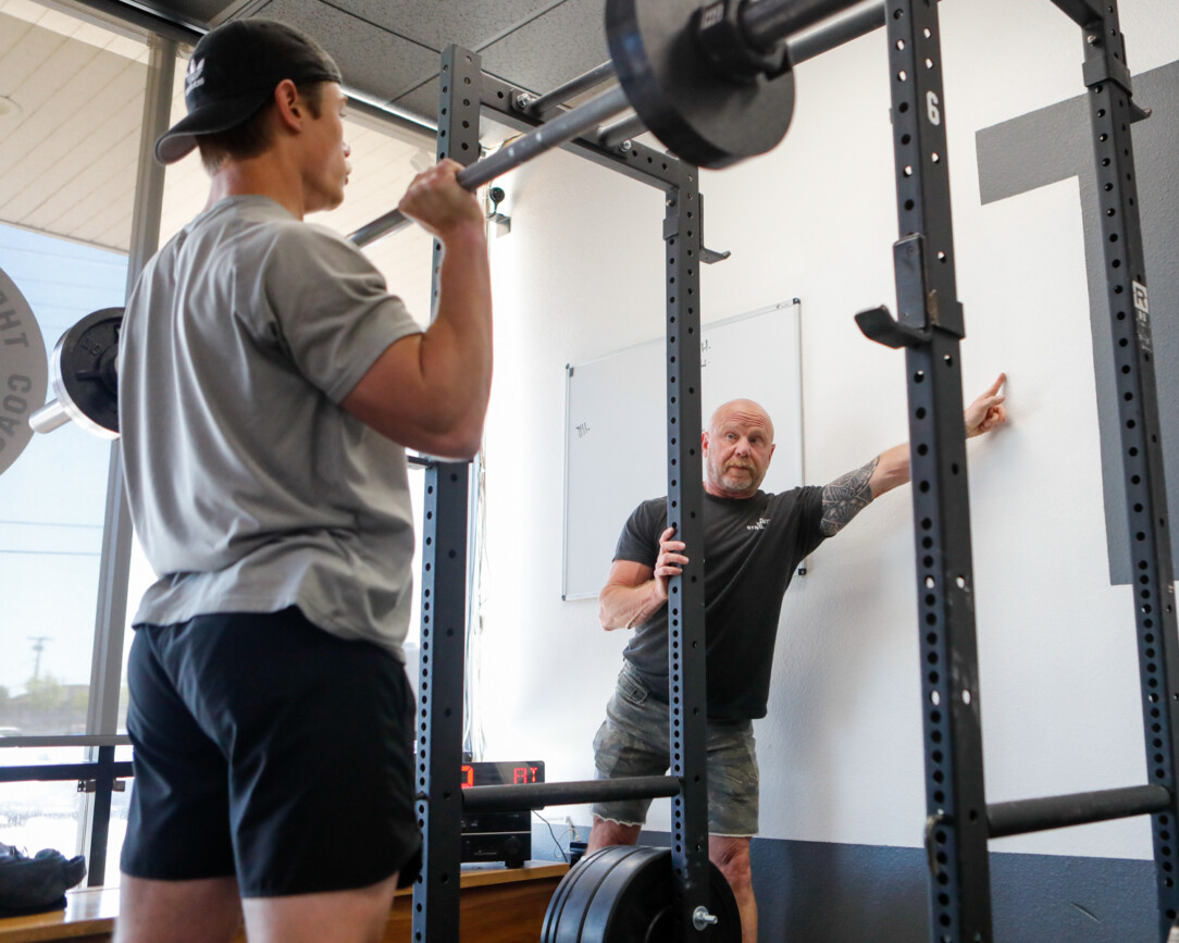 A man performs an overhead press while another man points at a spot on the wall to focus his attention.