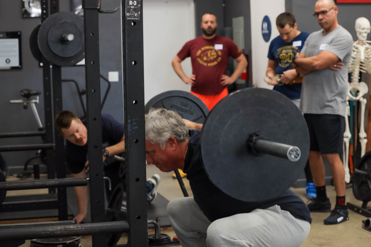 A man squats 145 pounds in a group class while a coach and other trainees look on.
