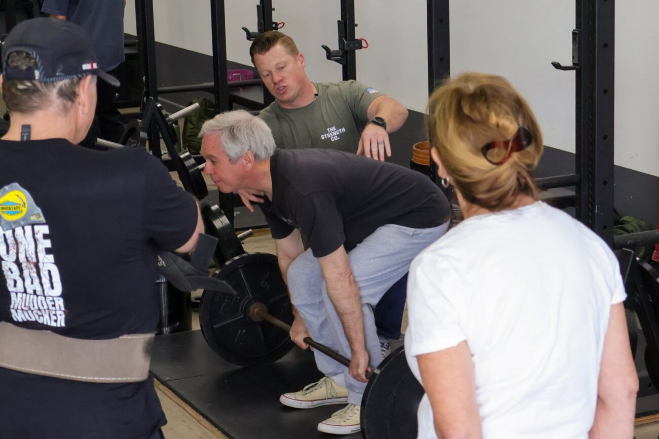 A man prepares to deadlift a barbell while a coach and trainees look on.