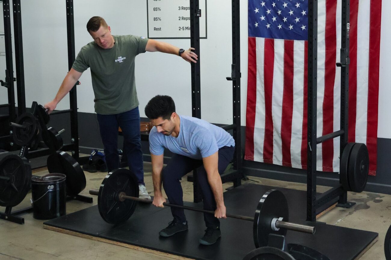 A man prepares to deadlift a barbell while a coach watches him.