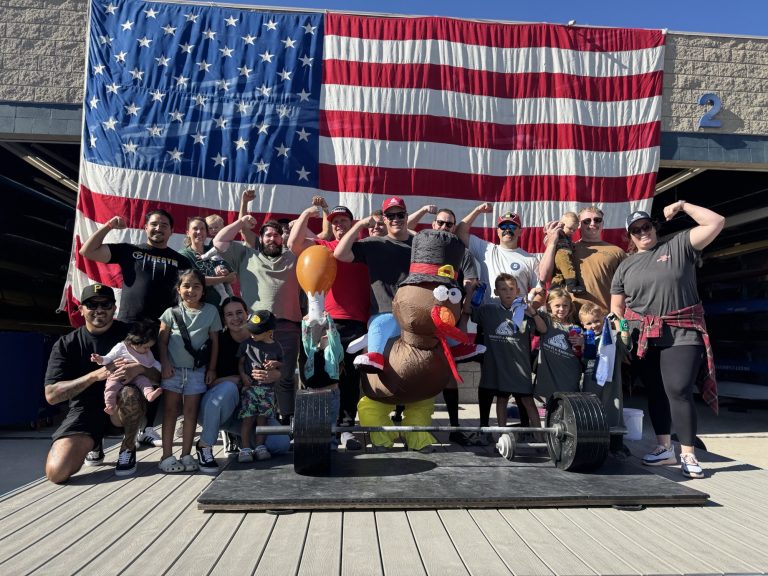 Lifters flexing behind a loaded barbell with the inflatable turkey mascot